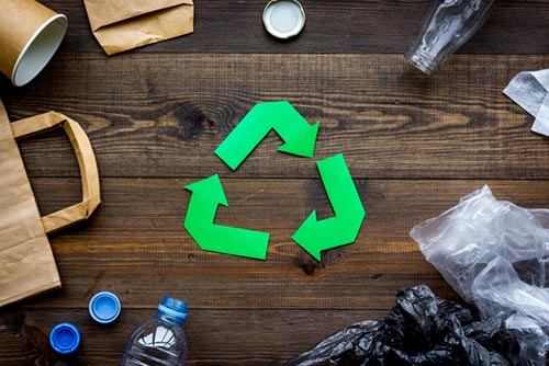 bottles bags and other recyclable material are placed on a wooden desk surrounding 3 green arrows that resemble the recycling logo